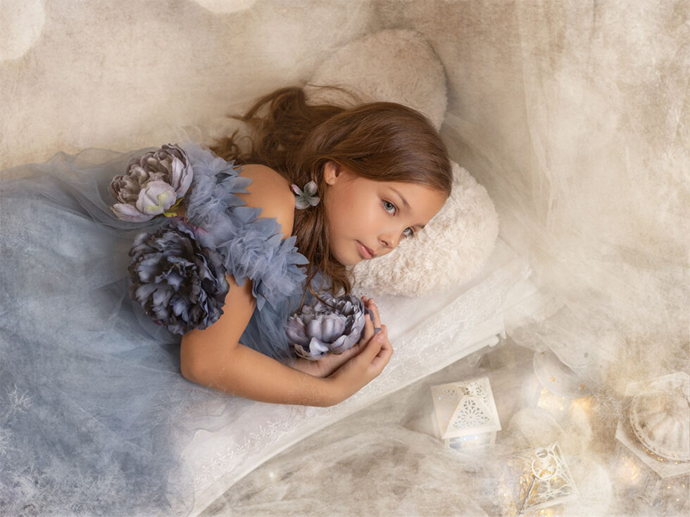 Fotografía niña vestido azul con flores acostada en cama fondo nubes blancas