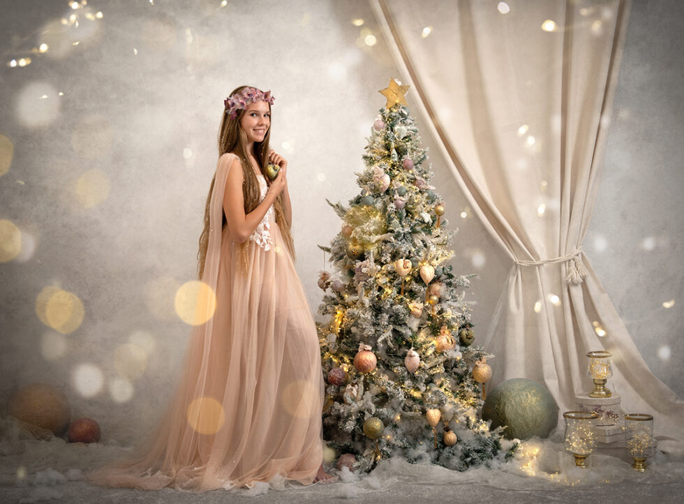 Fotografía de una chica con vestido rosa pálido al lado de un árbol de navidad en un fondo blanco