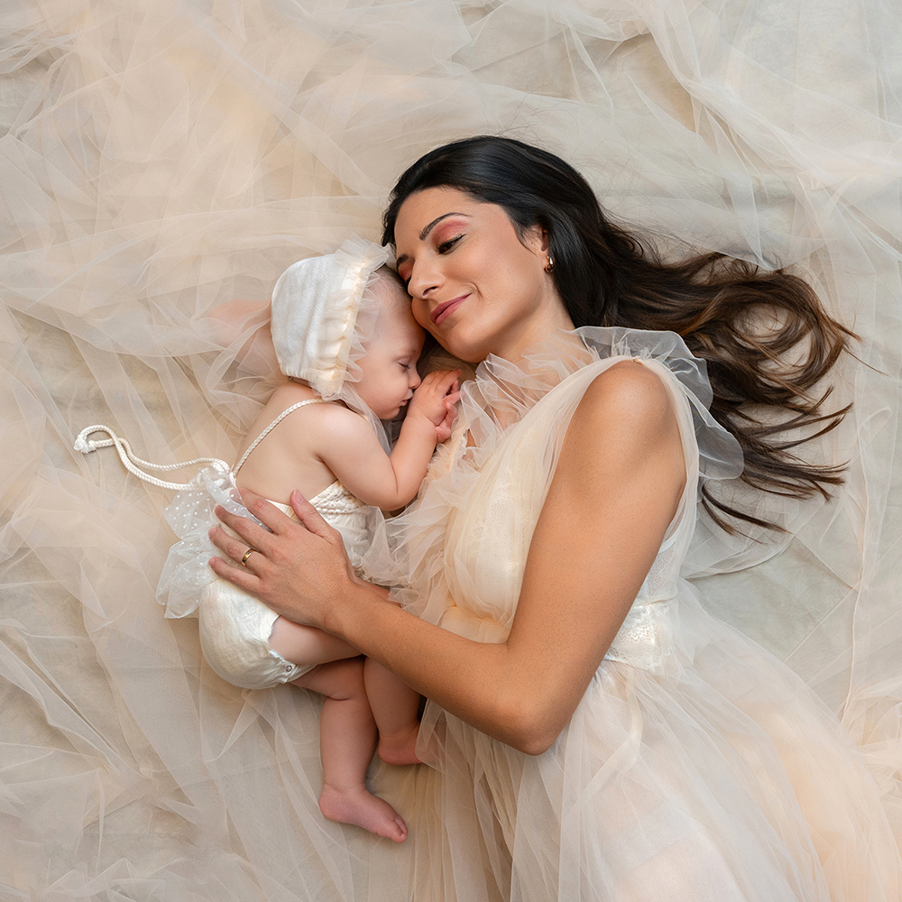Magicasfotos MamaYYo (16) Fotografía de una mujer con vestido blanco y su hija bebé acostadas en el suelo con vestido sonriendo a la cámara